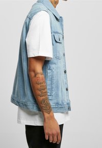 Světle modrá džínová vesta s límcem, předními knoflíky a dvěma kapsami na prsou; vrstvená přes bílé tričko.