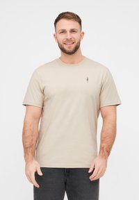 Beige T-Shirt mit kurzen Ärmeln aus Baumwolle. Mit einem kleinen dunkelbraunen Baum-Graphic auf der Brust. Rundhalsausschnitt und lässige Passform.