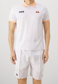 Homme portant un polo blanc avec un col pêche et un short blanc assorti, tous deux portant de petits logos de marque sur la poitrine et la jambe.