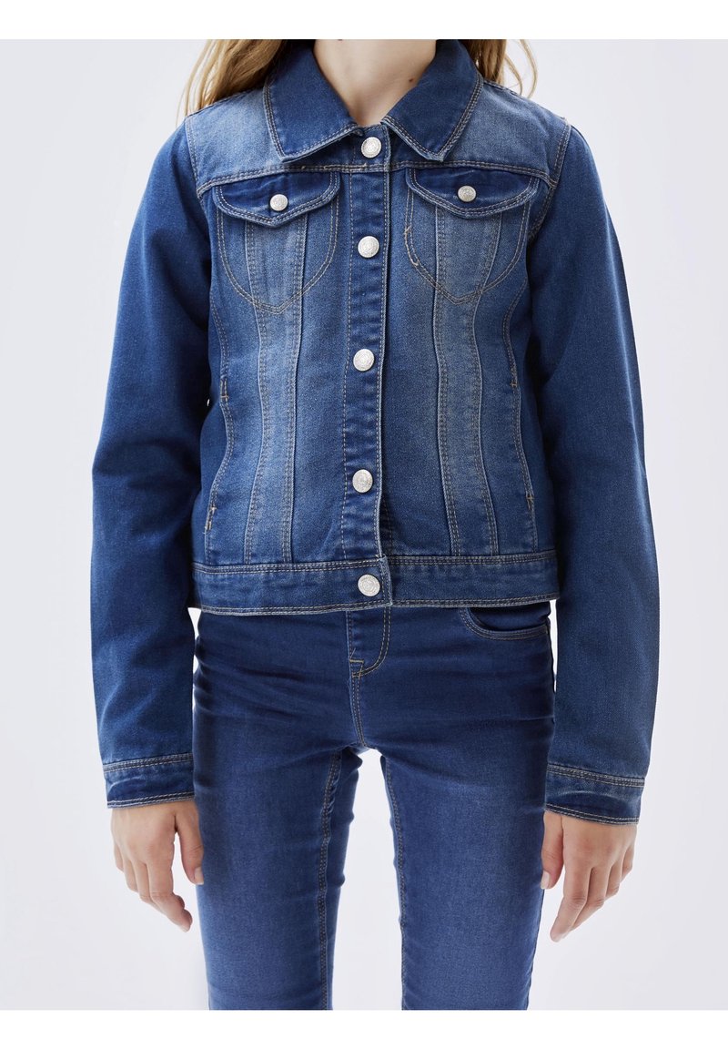 Enfant portant une veste en jean bleu ajustée avec des boutons argentés sur un jean en denim bleu assorti, debout devant un fond uni.