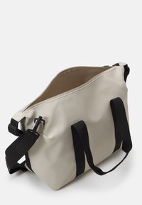 Beige vanntett totebag med svarte nylonstropper, med romslig innvendig plass og minimalistisk design. Inkluderer en sikker spenne-lukking.