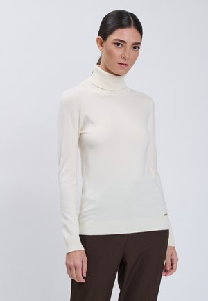 Femme aux cheveux foncés attachés en queue de cheval, vêtue d’un pull à col roulé blanc et d’un pantalon brun foncé, debout devant un fond clair uni.