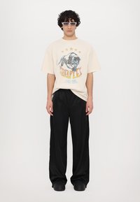 Beige oversized t-skjorte med grafikk av en panter og teksten "HOOPLES", kombinert med løse svarte bukser og kraftige svarte sko.