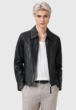 Veste en cuir noire avec un col classique, fermeture éclair et finition texturée. Portée sur une chemise blanche côtelée, complétée par un pantalon léger.