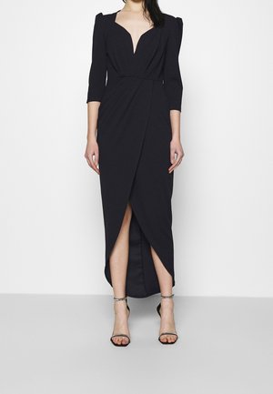 Zwarte aansluitende jurk met een diepe V-hals, driekwart mouwen en een zijsplit, gemaakt van gladde stof. Geaccentueerd met strakke hakken.