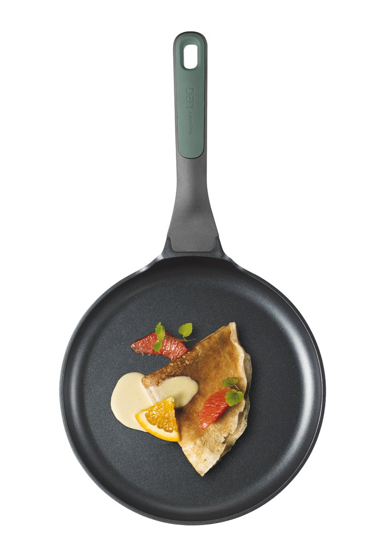Schwarze antihaftbeschichtete Pfanne mit grünem Griff, gefüllt mit einem Pfannkuchen, der mit Sahne, orangefarbenen und roten Früchten sowie Minzblättern garniert ist.