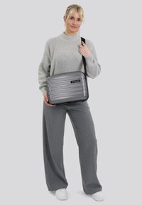 Borsa a tracolla rigida grigia con scanalature orizzontali, un logo nero e una tracolla regolabile, tenuta da una persona in abbigliamento grigio.