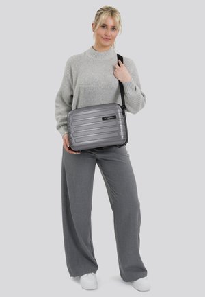 Grijze harde schoudertas met horizontale ribbels, een zwart logo en een verstelbare strap, vastgehouden door een persoon in grijze kleding.