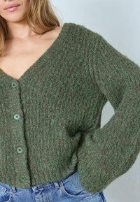Zaļš adīts kardigans ar V-veida kakla izgriezumu, strukturētu rakstu un lielajiem pogām. Iespējas: garas, pūkainas piedurknes un brīva piegriezuma siluets.