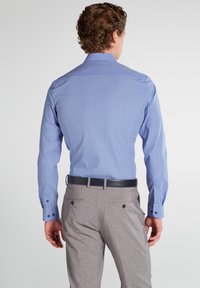 Man with curly hair wearing a light blue dress shirt tucked into gray trousers with a black belt, facing away against a plain background.