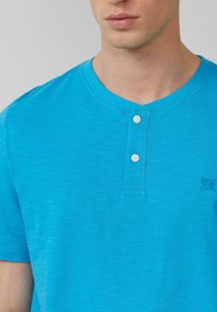 Helles türkisfarbenes T-Shirt mit kurzen Ärmeln und rundem Halsausschnitt, versehen mit zwei weißen Knöpfen und einem kleinen Logo auf der linken Seite.