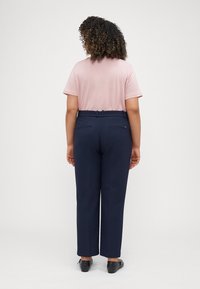 Marineblaue weit geschnittene Hose mit Gesäßtaschen, kombiniert mit einem hellrosa T-Shirt mit kurzen Ärmeln. Das Model steht mit dem Rücken zur Kamera und zeigt die Passform des Outfits.