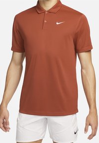 Homme portant un polo Nike couleur rouille et un short Nike blanc avec logos visibles, debout les bras détendus le long du corps.