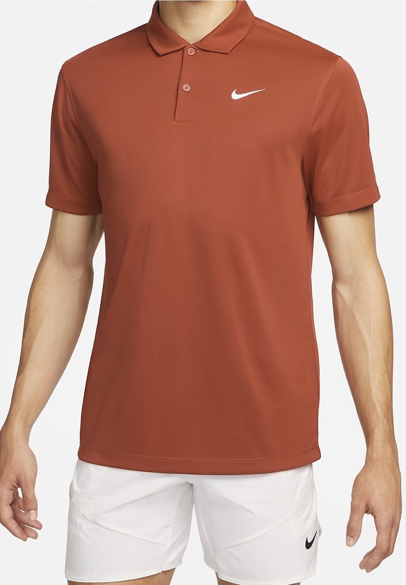 Homme portant un polo Nike couleur rouille et un short Nike blanc avec logos visibles, debout les bras détendus le long du corps.