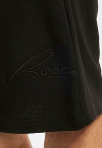 Black athletic shorts made of soft fabric, featuring an embroidered "Rocawear" logo in a matching color along the side seam and a simple cut.
