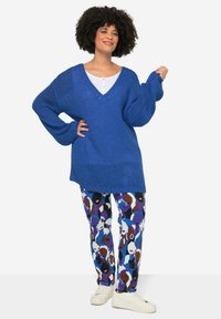Blauw oversized gebreid sweater met een V-hals en lange mouwen, gecombineerd met gestreepte broeken met kleurrijke abstracte gezichten op een witte achtergrond.
