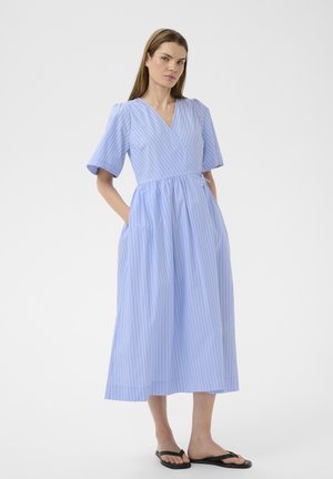 Woman wearing a light blue, white-striped midi wrap dress with short sleeves, standing with hands in pockets, and black flip-flops on white background.