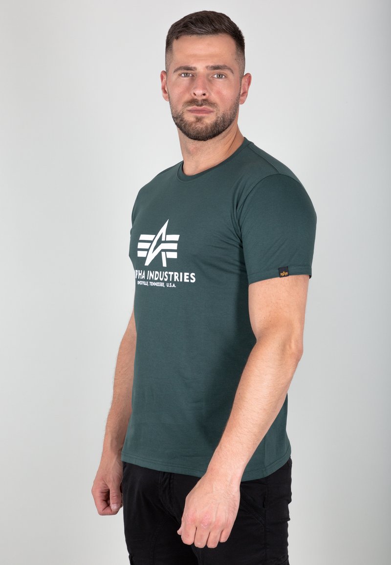 Grünes T-Shirt mit kurzen Ärmeln aus Baumwolle, mit einem weißen Logo "Alpha Industries" auf der Vorderseite und einem schwarzen Akzentlabel am Ärmel.