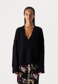 Schwarzer, übergroßer Pullover mit tiefem V-Ausschnitt und strukturiertem Stoff. Weiche, locker sitzende Ärmel. Kombiniert mit blumengemusterten Hosen.