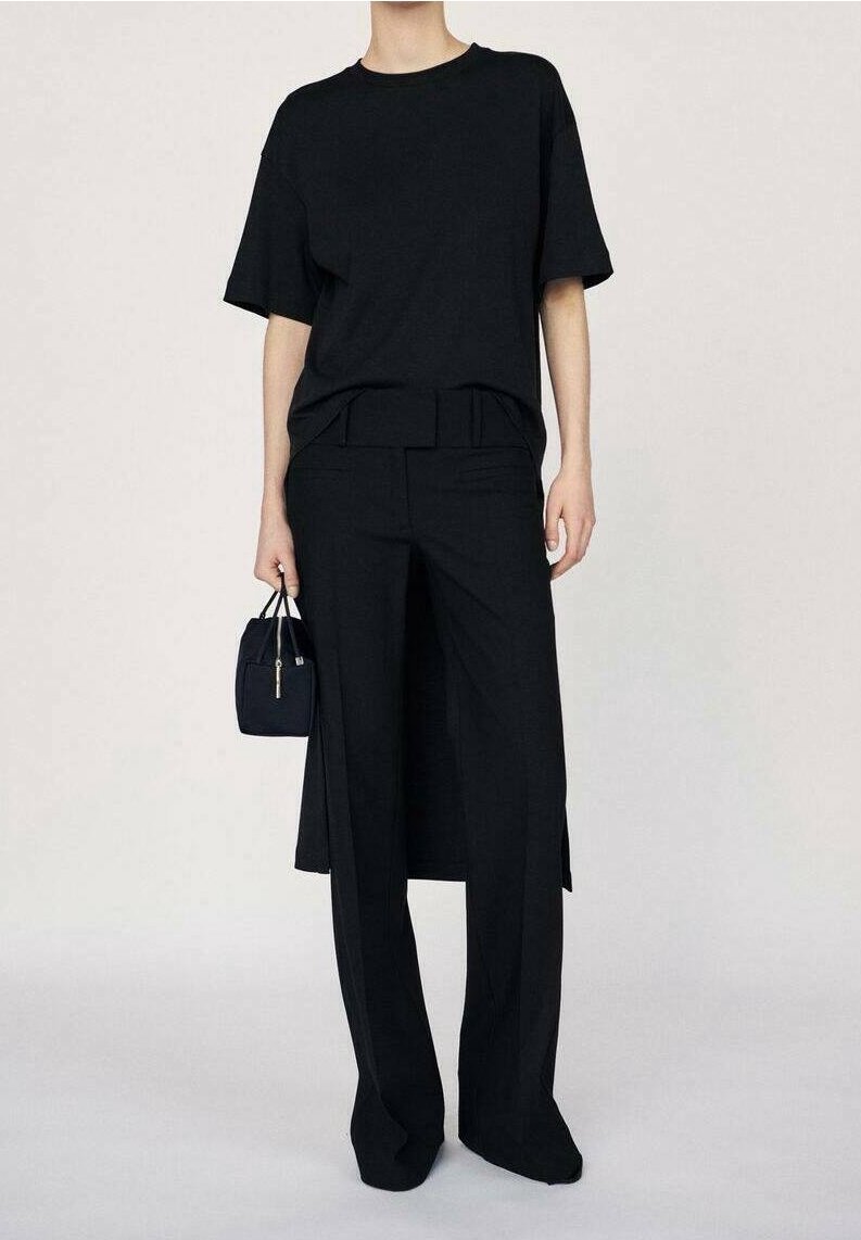 Personne portant un t-shirt noir ample, un pantalon noir à jambes larges, tenant un petit sac à main noir sur un fond clair uni.