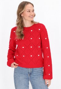 Pulover tricotat roșu cu un model de diamant și accente albe în formă de inimă. Guler și mansete cu eleastan. Asortat cu blugi din denim albastru.