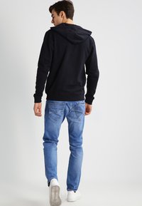 Sweatshirt à capuche noir, coupe décontractée, associé à un jean bleu clair. Le modèle porte des baskets blanches, vu de dos.