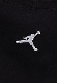 Crna tkanina s bijelim izvezenim Jumpman logotipom, koji prikazuje figuru u sportskom stavu s podignutom rukom, na glatkoj teksturi.