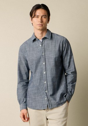 Junger Mann, der ein blaues Hemd mit langen Ärmeln und beige Hose trägt und vor einem einfarbigen beigen Hintergrund steht.