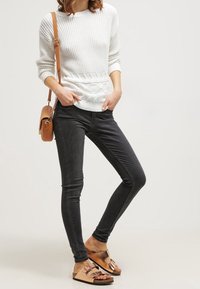 Biały prążkowany sweter z teksturowanym paskiem w talii, zestawiony z czarnymi obcisłymi dżinsami i brązowymi sandałami z metalicznymi akcentami i paseczkami.