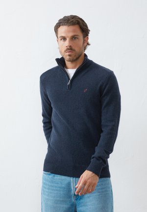 Navy zip-up sweater in soft fabric with ribbed cuffs and hem. Small embroidered logo on chest. Worn over a white T-shirt and paired with light jeans.