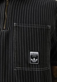 Mustriline mustri särk, millel on esikülg tasku, pronksist vetti ja Adidas logo plaaster. Materjalil on koobasaine tekstuur ja kontrastne õmblus.