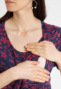 Laivansinis mekko, jossa on burgundinvärinen kukkakuosi. Naisen kädet lepäävät rinnan alueella, ja mekossa on vaalean sävyinen lisäosa. Suuret renkaat korvissa.