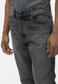 Close-up of a person wearing dark gray denim jeans with front pockets and a buttoned waistband, standing against a white background.