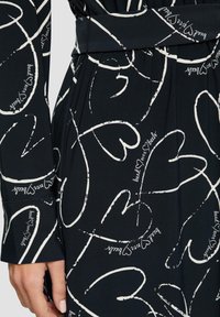 Tessuto nero con contorni di cuori bianchi e motivi di testo scritto a mano. Presenta una texture liscia e un design con vita elastica.