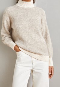 Beiger flauschiger Pullover mit hohem Rippkragen und cremefarbenen Ärmeln, im lässigen Schnitt. Kombiniert mit weißen Hosen, die eine weiche Textur zeigen.