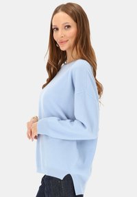 Jasnoniebieski sweter z miękkiej dzianiny, o luźnym kroju, z szerokim dekoltem i rozcięciami po bokach przy dolnym brzegu. Noszony na ciemnych dżinsach.