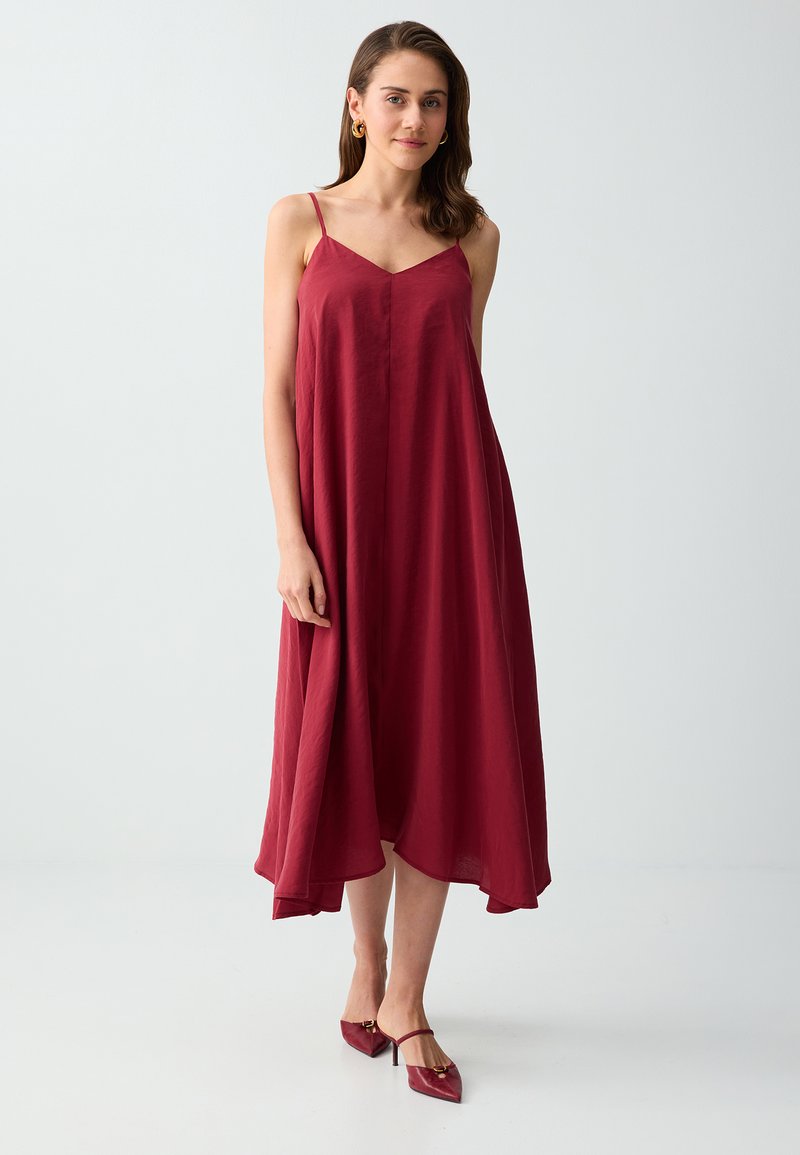 Femme debout, vêtue d'une robe midi bordeaux sans manches avec fines bretelles et des escarpins pointus assortis, sur un fond clair uni.