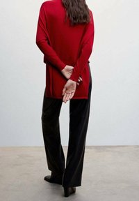Pull en tricot rouge avec une coupe ample, associé à un pantalon large noir. L'arrière du pull présente un ourlet asymétrique. Des bottes et des mains sont visibles à l'arrière.