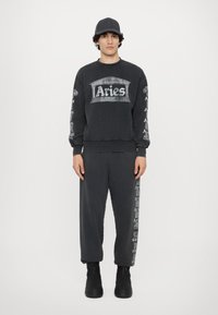 Sort sweatshirt med "Aries"-logo og søjlegrafik på brystet, parret med matchende bukser med logo på siden. Båret med en sort kasket.
