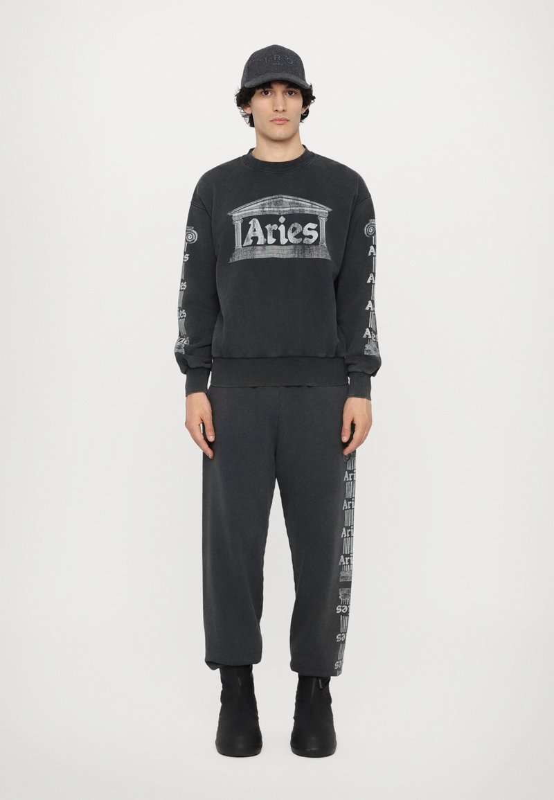 Sort sweatshirt med "Aries"-logo og søjlegrafik på brystet, parret med matchende bukser med logo på siden. Båret med en sort kasket.
