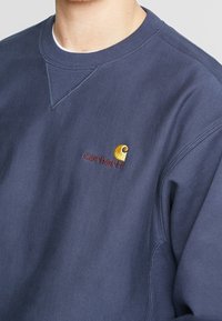 Man i marinblå Carhartt-tröja med broderad logga i gult och brunt på bröstet.