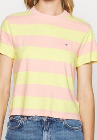 Mujer con una camiseta de manga corta en tonos rosa y amarillo a rayas, con un logo en el pecho, y unos vaqueros de cintura alta en color azul.