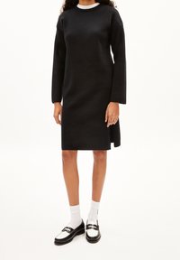 Robe en maille noire à manches longues, col rond et fente latérale. Portée avec des chaussettes blanches et des chaussures noires et blanches. Texture lisse.