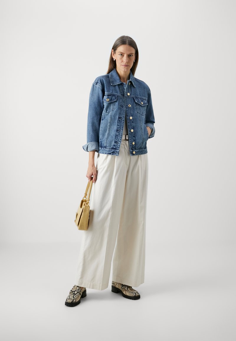 Chaqueta de mezclilla en azul medio, con cierres de botones y dos bolsillos en el pecho, llevada sobre pantalones de pierna ancha en beige claro y zapatos con estampado.