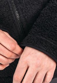 Detail einer schwarzen Fleecejacke, die einen strukturierten Stoff zeigt, mit einer Nahaufnahme eines Ärmels, der einen glatten schwarzen Besatz und einen Reißverschluss entlang der Kante aufweist.