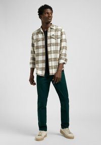 Camicia di flanella a scacchi in crema, marrone e nero, abbinata a pantaloni verde scuro e sneakers beige. Due tasche sulla camicia.