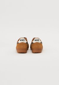 Tênis Nike com parte superior em camurça bege, detalhes texturizados em preto e um tabuleiro branco no calcanhar com o logótipo da marca. Sola de borracha plana com aderência.