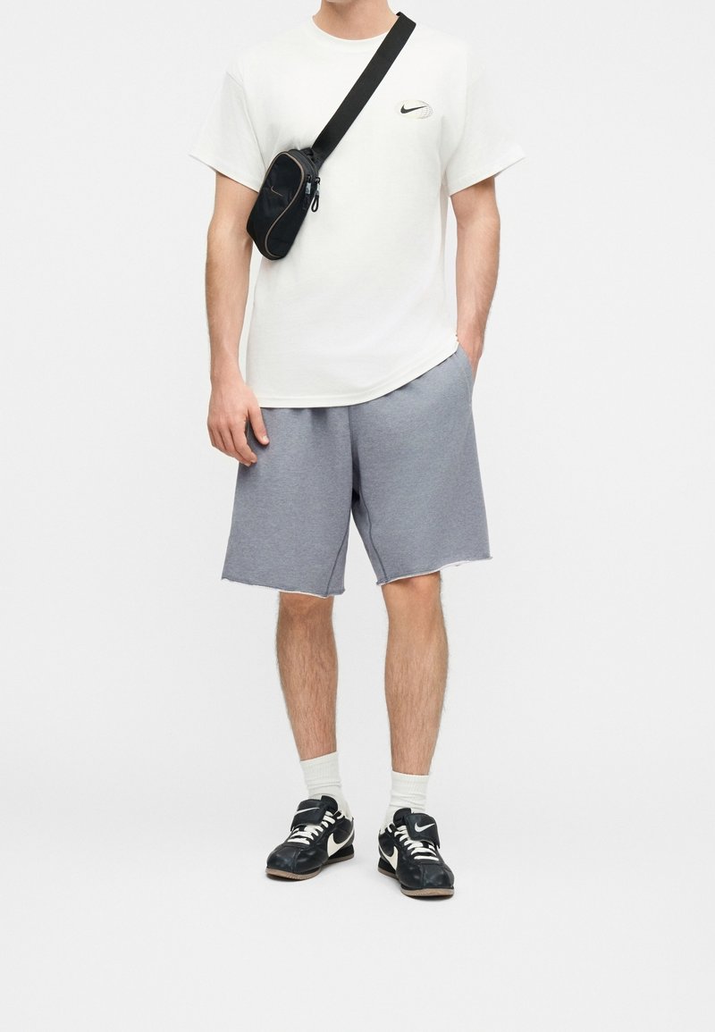 Homme portant un t-shirt blanc Nike, un short gris, un sac bandoulière noir, des chaussettes blanches et des baskets Nike noires, debout les mains dans les poches.