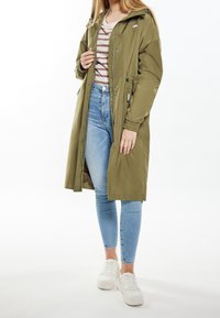 Green, water-resistant long jacket with a hood, elastic cuffs, and buttons. Paired with a striped shirt and jeans, completed with white sneakers.
