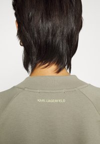 Skrót w kolorze oliwkowym z okrągłym dekoltem, wyróżniający się wyraźną struktura prążkową oraz haftem "KARL LAGERFELD" w jasnym zielonym na plecach.